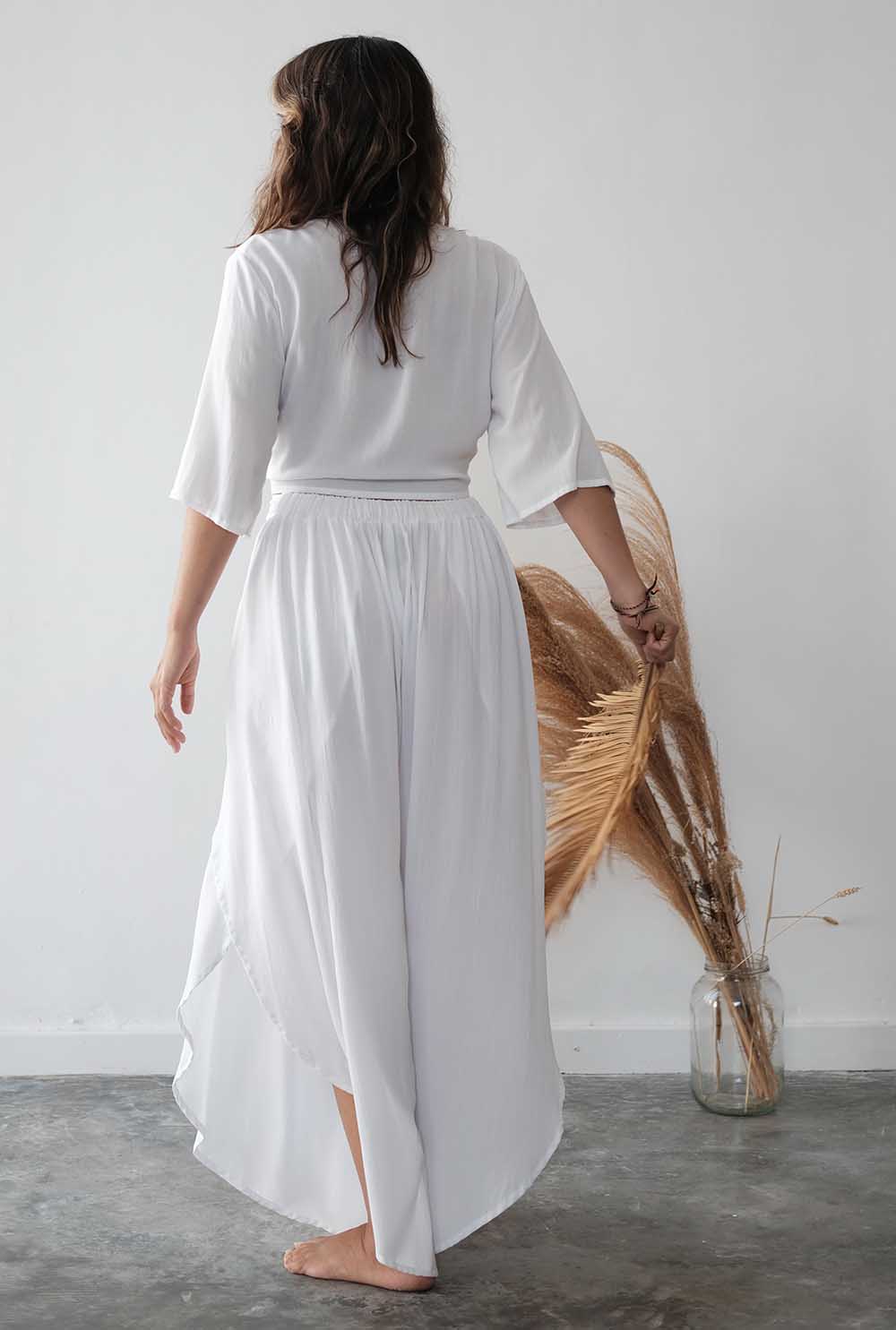 Woman wearing a white outfit standing in a minimalistic room with a vase and dried plant.