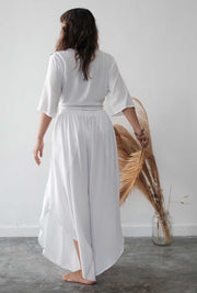 Woman wearing a white outfit standing in a minimalistic room with a vase and dried plant.