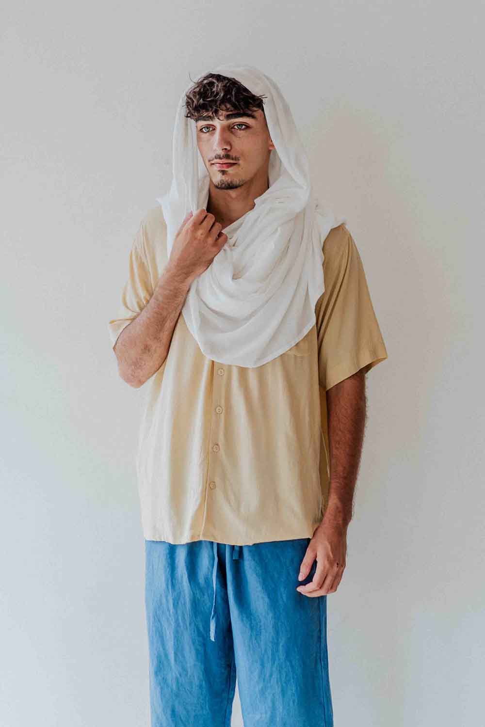 Person wearing a beige shirt and blue pants with a white scarf on a plain background