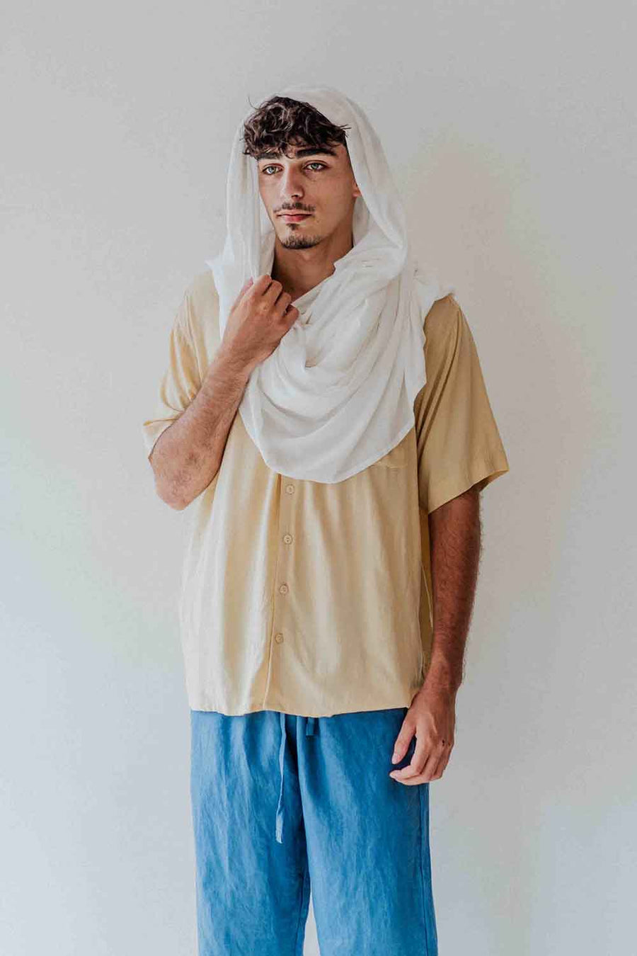 Person wearing a beige shirt and blue pants with a white scarf on a plain background