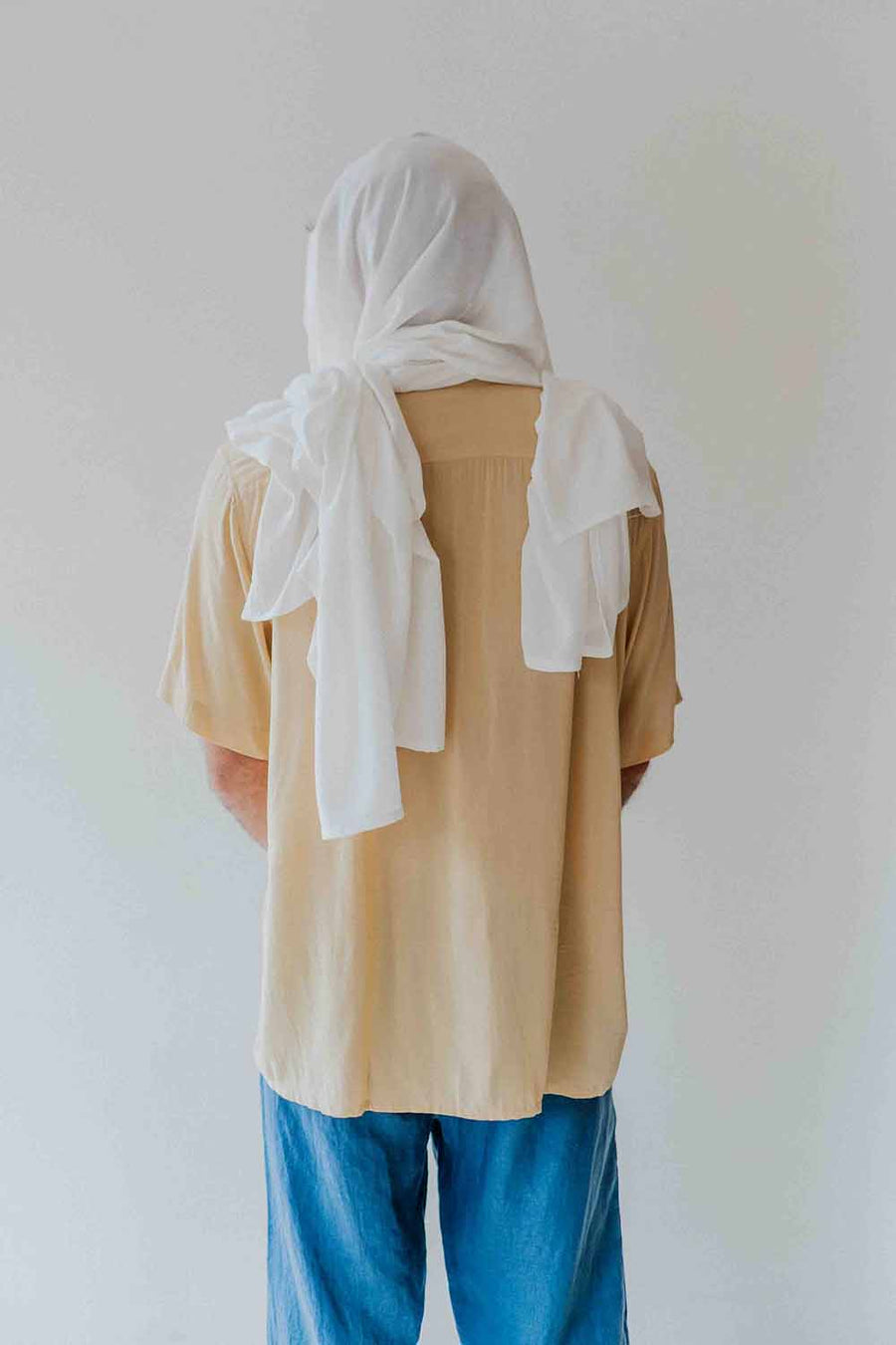 Person wearing a beige shirt and blue pants with a white scarf on a plain background