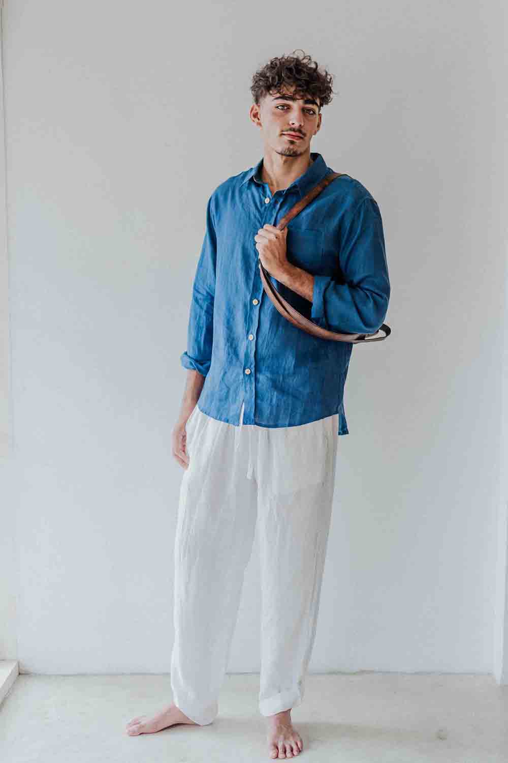 Man wearing a blue shirt and white pants holding a brown leather bag against a white background