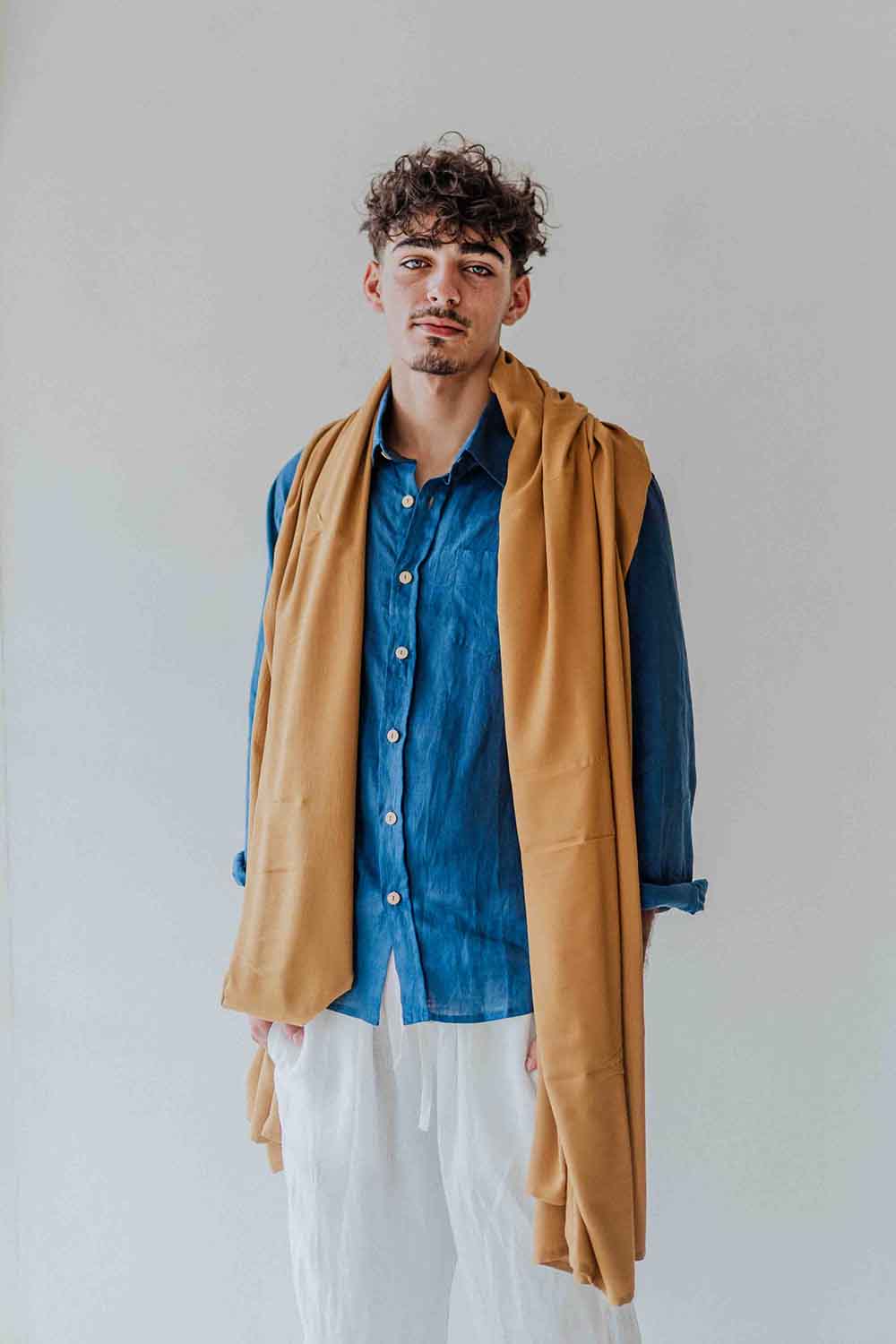 Man wearing a blue shirt, white pants, and a mustard yellow jacket against a plain background