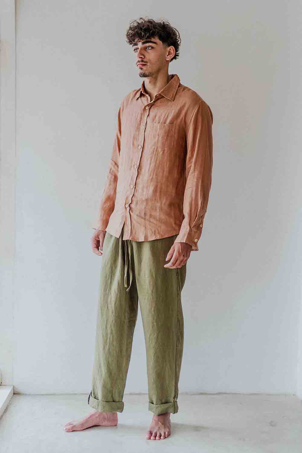 Man wearing a light brown shirt and green pants standing against a white wall.