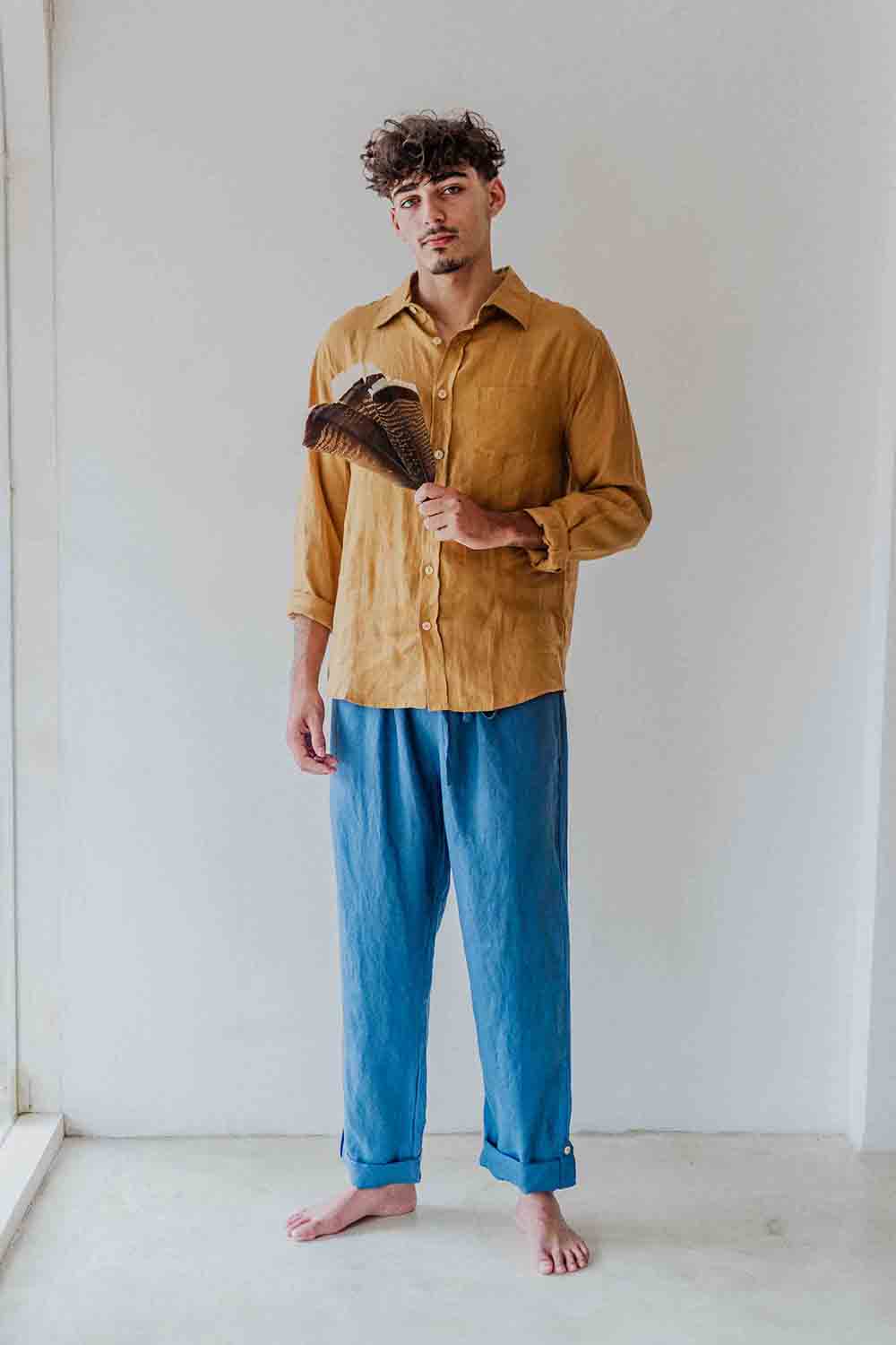 Man wearing a mustard yellow shirt and blue pants holding a feather duster against a white wall.