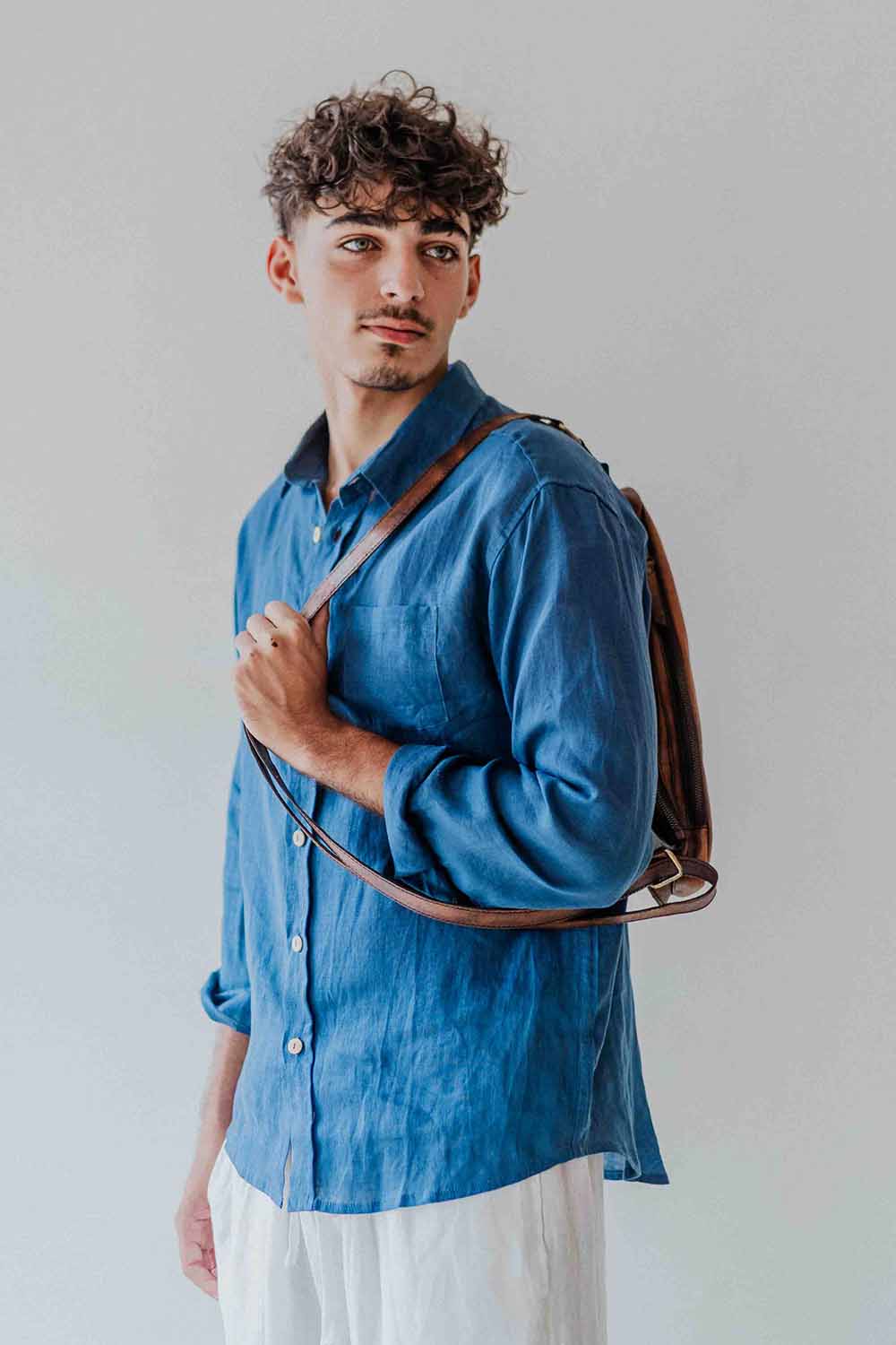 Man wearing a blue shirt holding a brown leather bag against a plain background