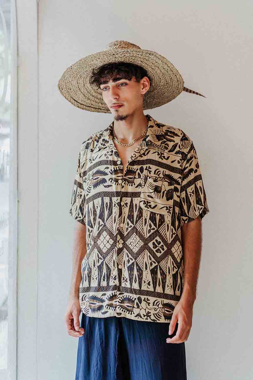 Man wearing a patterned shirt and straw hat against a white wall