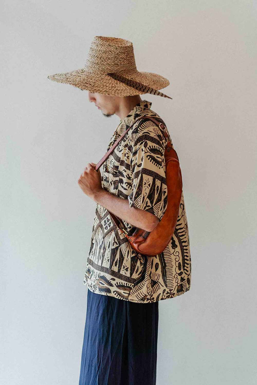 Person wearing a patterned shirt and straw hat with a brown bag against a white background