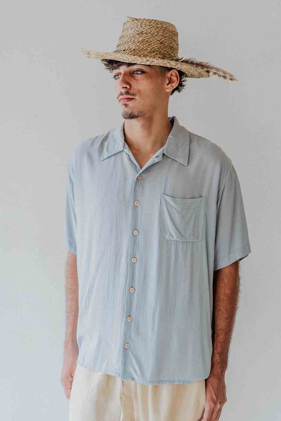 Man wearing a light blue button-up shirt and straw hat against a plain background