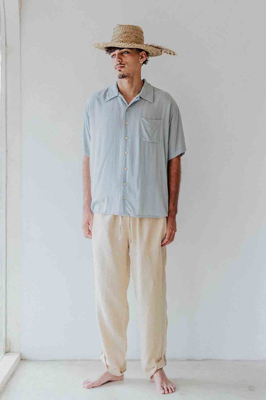 Man wearing a light blue shirt and beige pants with a straw hat against a white background