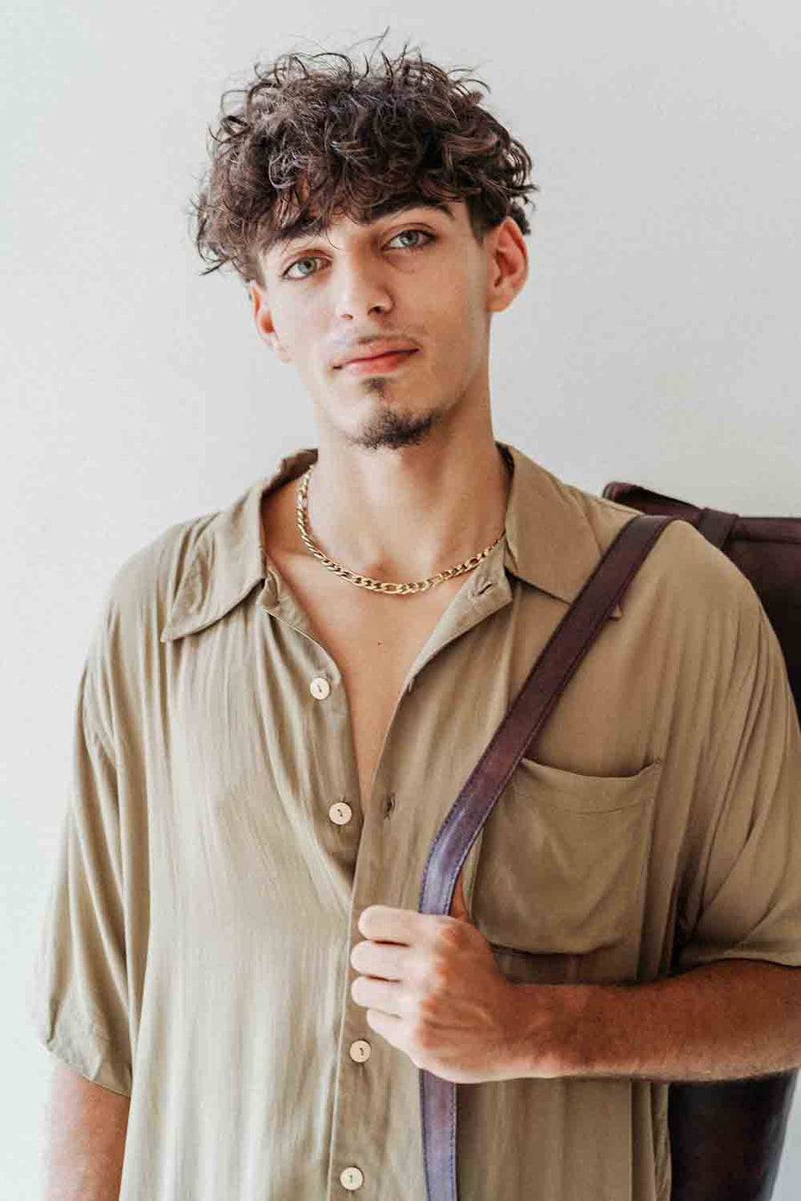 Man wearing a beige shirt with a brown strap over his shoulder against a light background