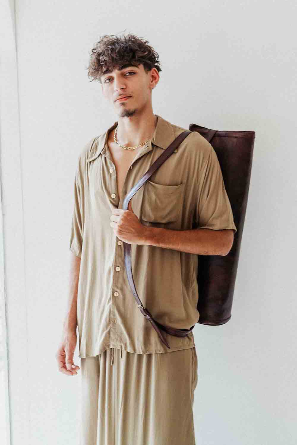 Man wearing a beige shirt and pants holding a brown leather bag against a white background