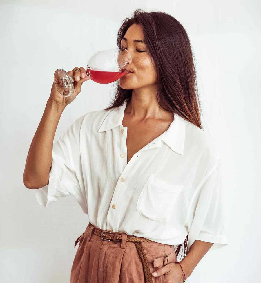 Woman in a white shirt and brown pants drinking from a glass of red wine against a white background