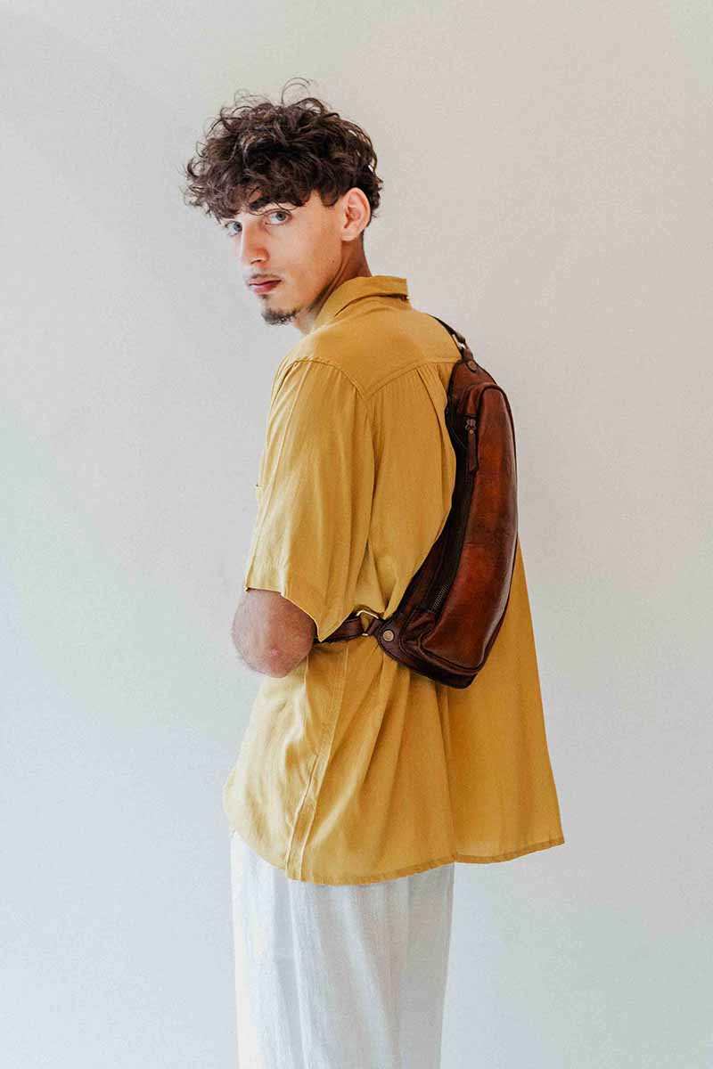 Man wearing a mustard yellow shirt with a brown leather bag over his shoulder against a white background