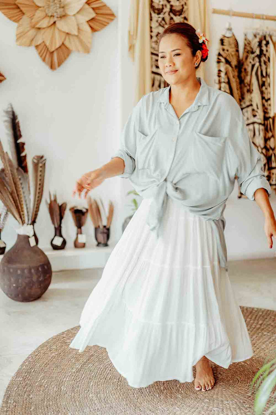 Woman in a light blue shirt and white skirt standing in a room with decorative items on the wall.