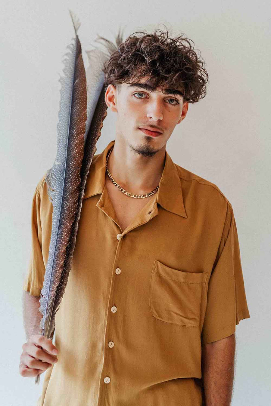 Man in a mustard yellow shirt holding a large feather against a plain background
