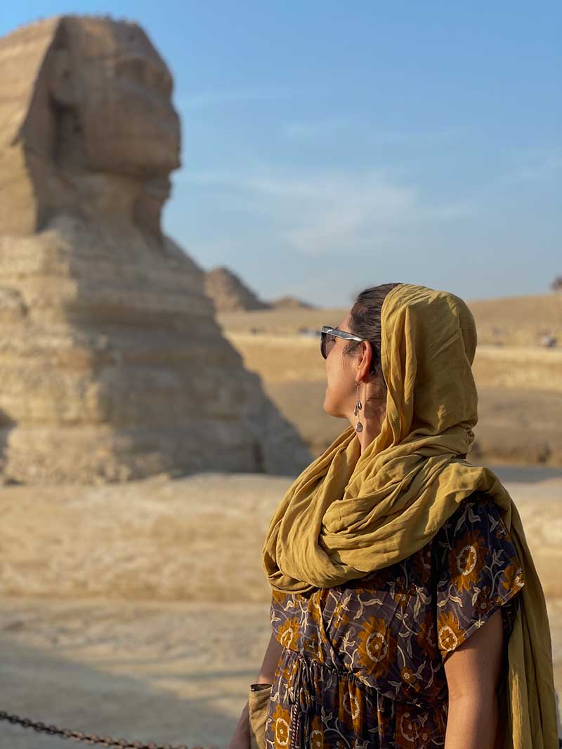 Woman in a patterned dress and yellow headscarf standing in front of the Sphinx at the Giza Plateau.