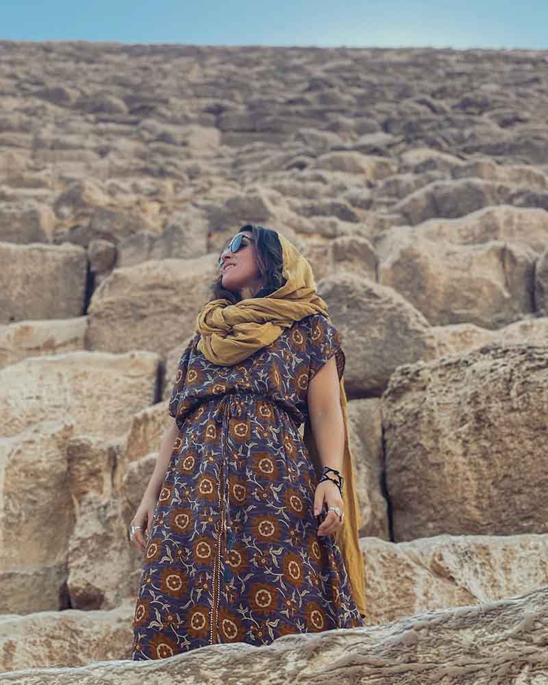 Woman in a patterned dress and yellow scarf standing among stone ruins.