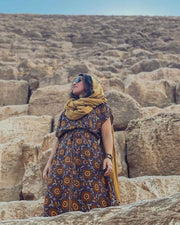 Woman in a patterned dress and yellow scarf standing among stone ruins.