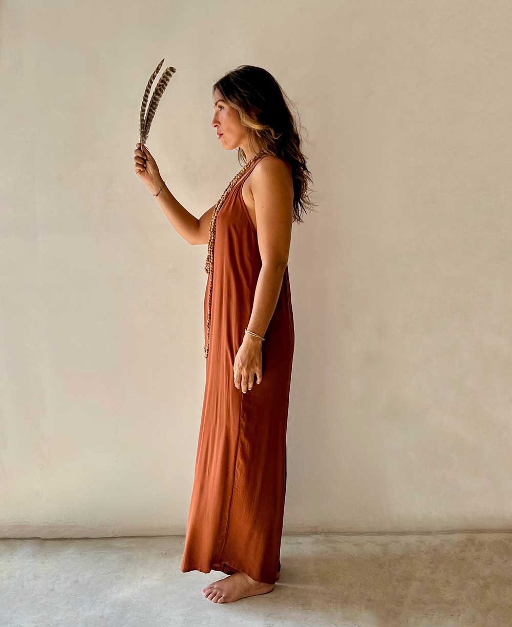 Woman in a rust-colored dress holding feathers against a beige wall