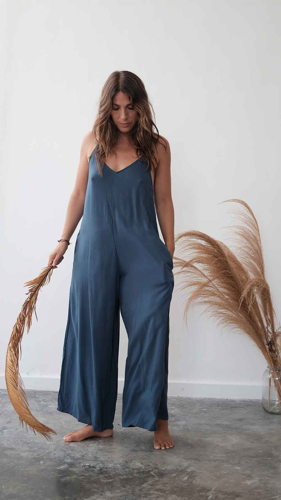 Woman wearing a blue jumpsuit standing against a white wall with dried plants.