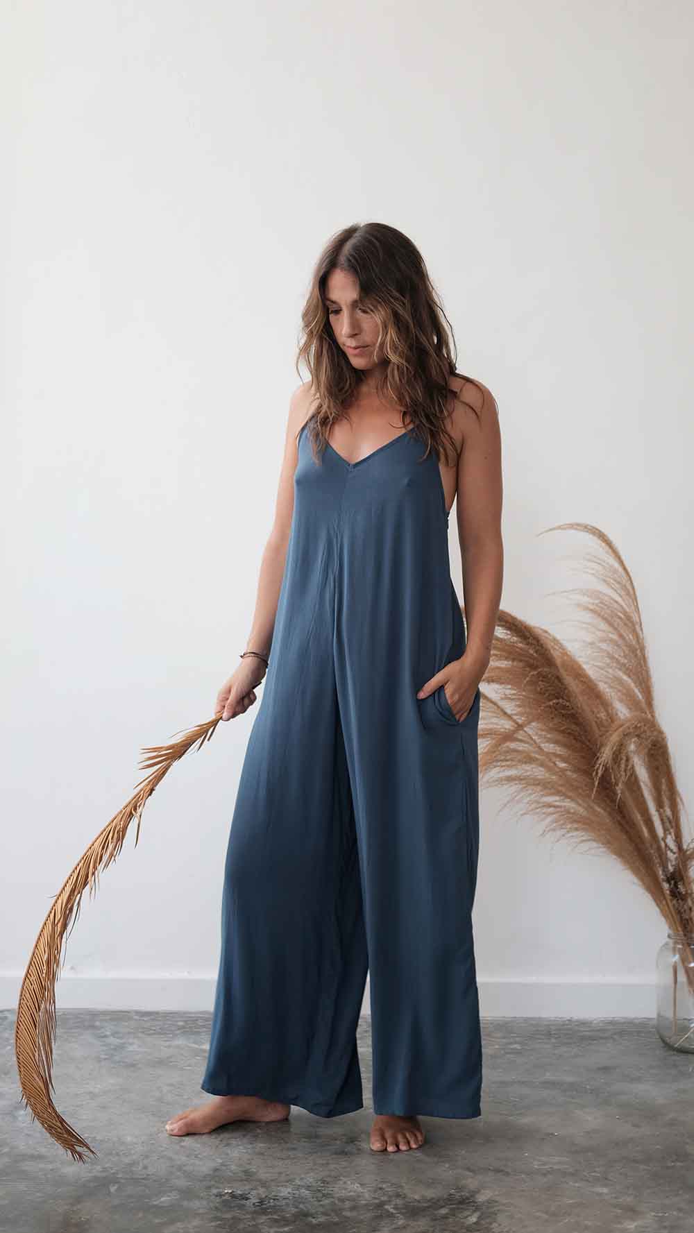 Woman wearing a blue jumpsuit standing against a white wall with dried pampas grass.