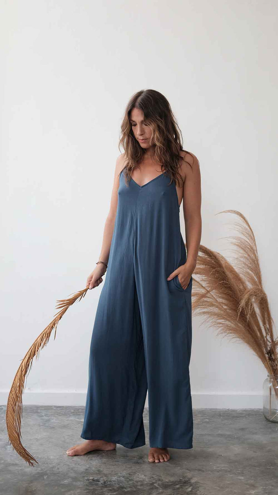 Woman wearing a blue jumpsuit standing against a white wall with dried pampas grass.