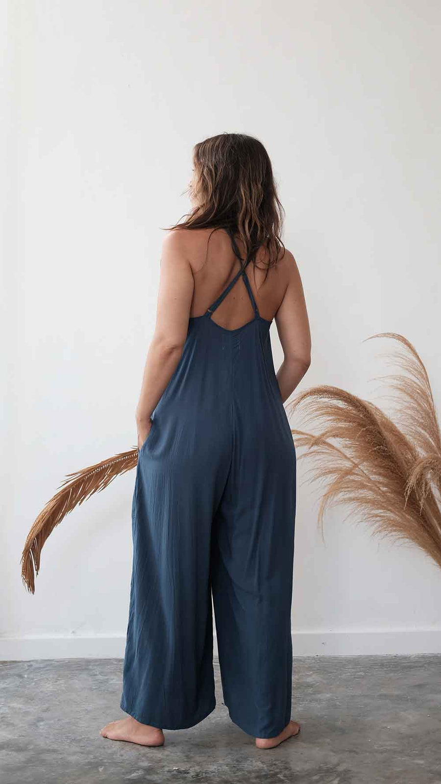 Woman wearing a blue jumpsuit standing against a white wall with dried plants.