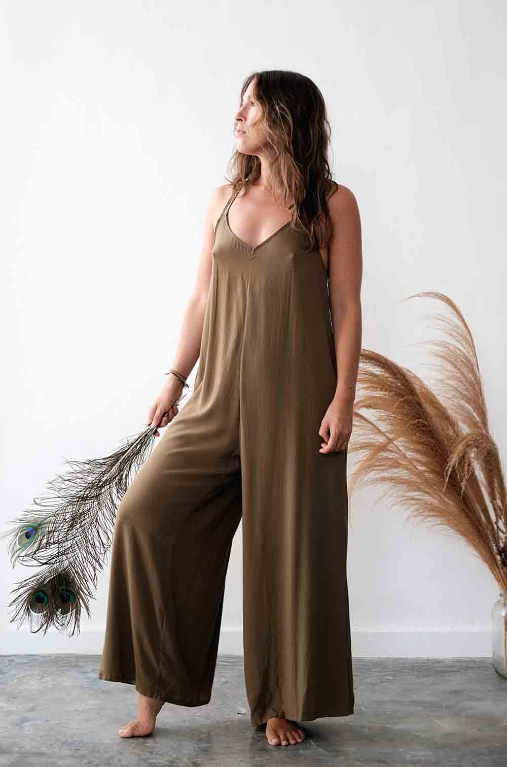 Woman wearing a brown jumpsuit holding dried plants against a white wall.