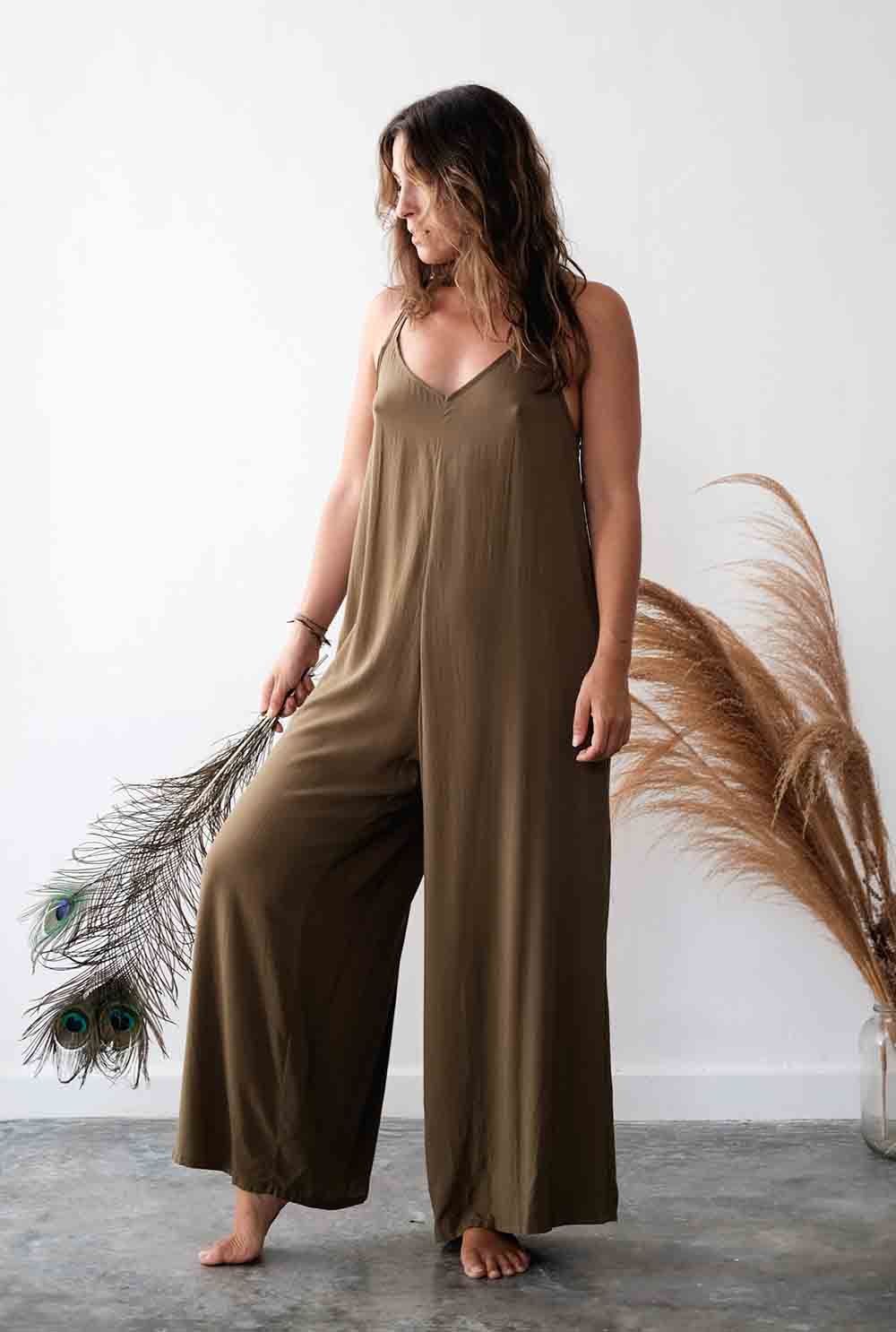 Woman wearing a brown jumpsuit standing next to dried pampas grass against a white wall.