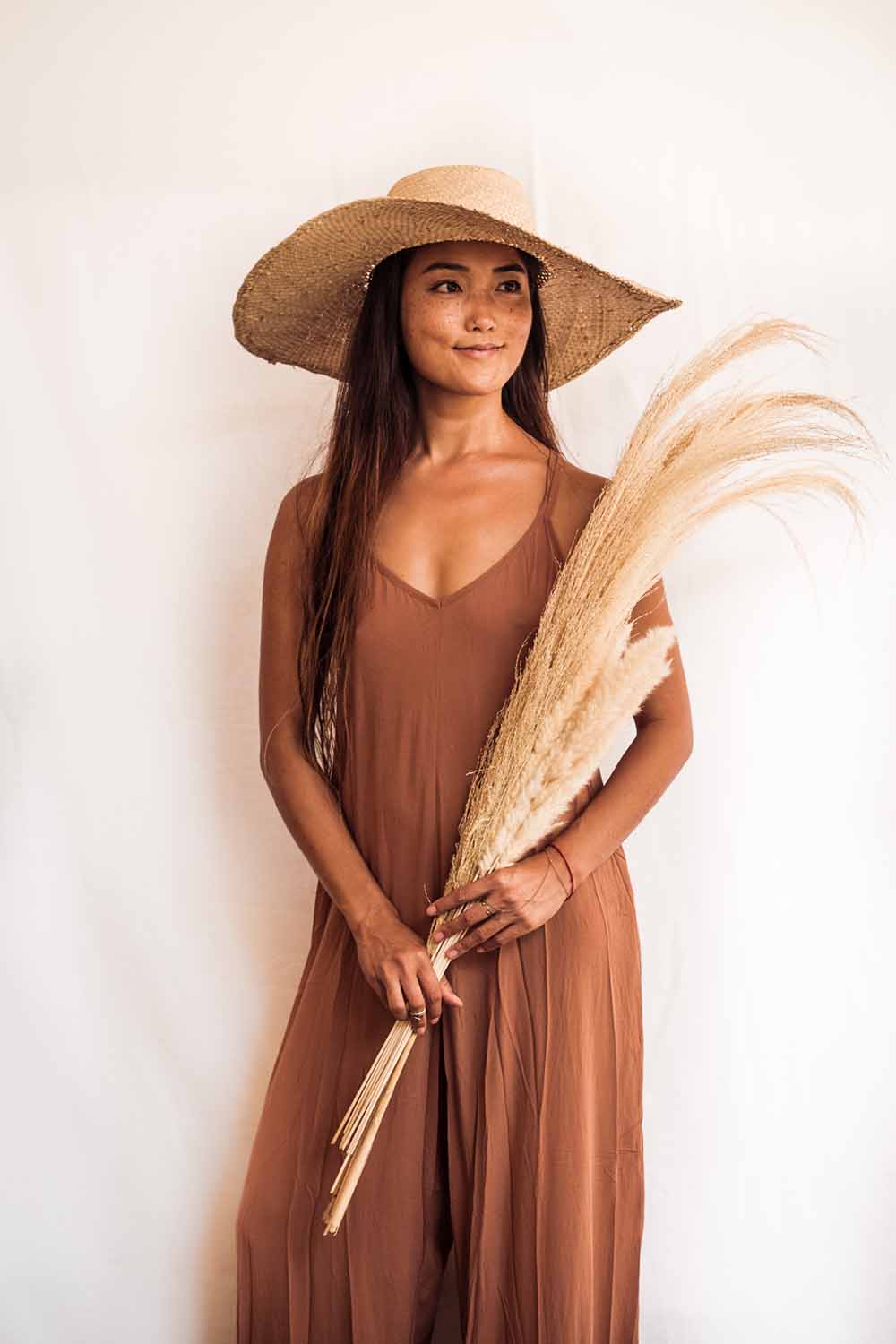 Woman in a brown dress and hat holding dried plants against a white background
