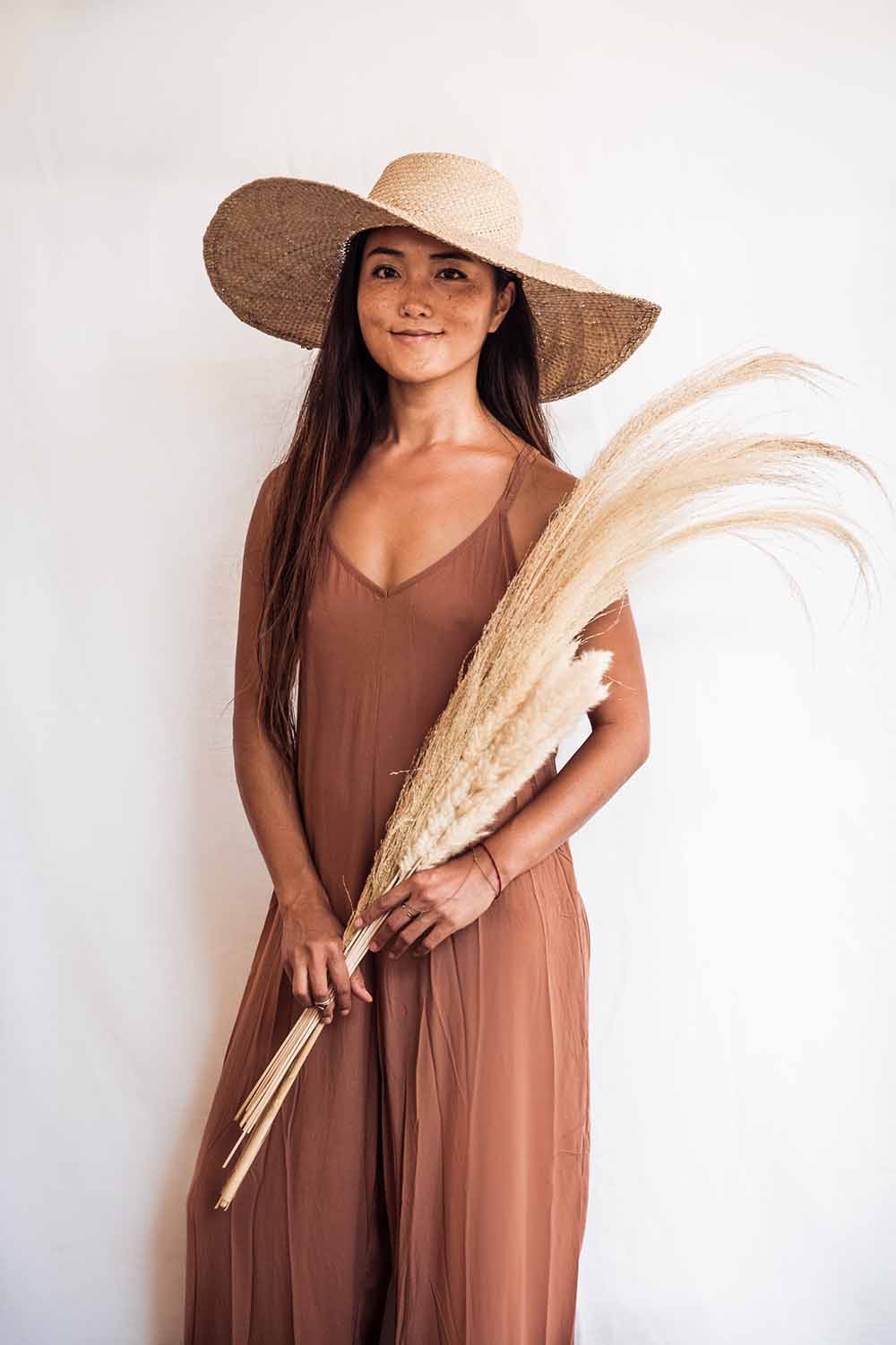 Woman in a long brown dress and wide-brimmed hat holding dried plants against a white background