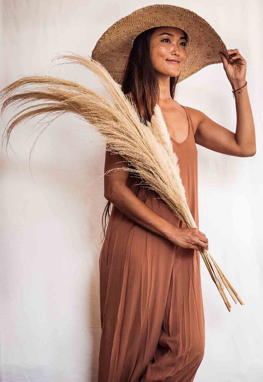 Woman in a brown dress and straw hat holding pampas grass against a white background