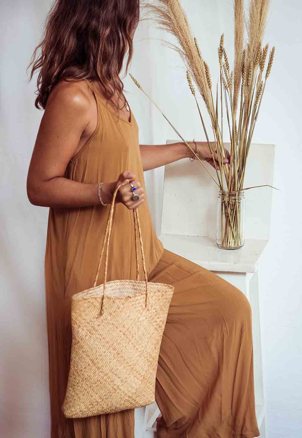 Woman in a mustard dress holding a woven bag with a vase of dried plants on a white surface.