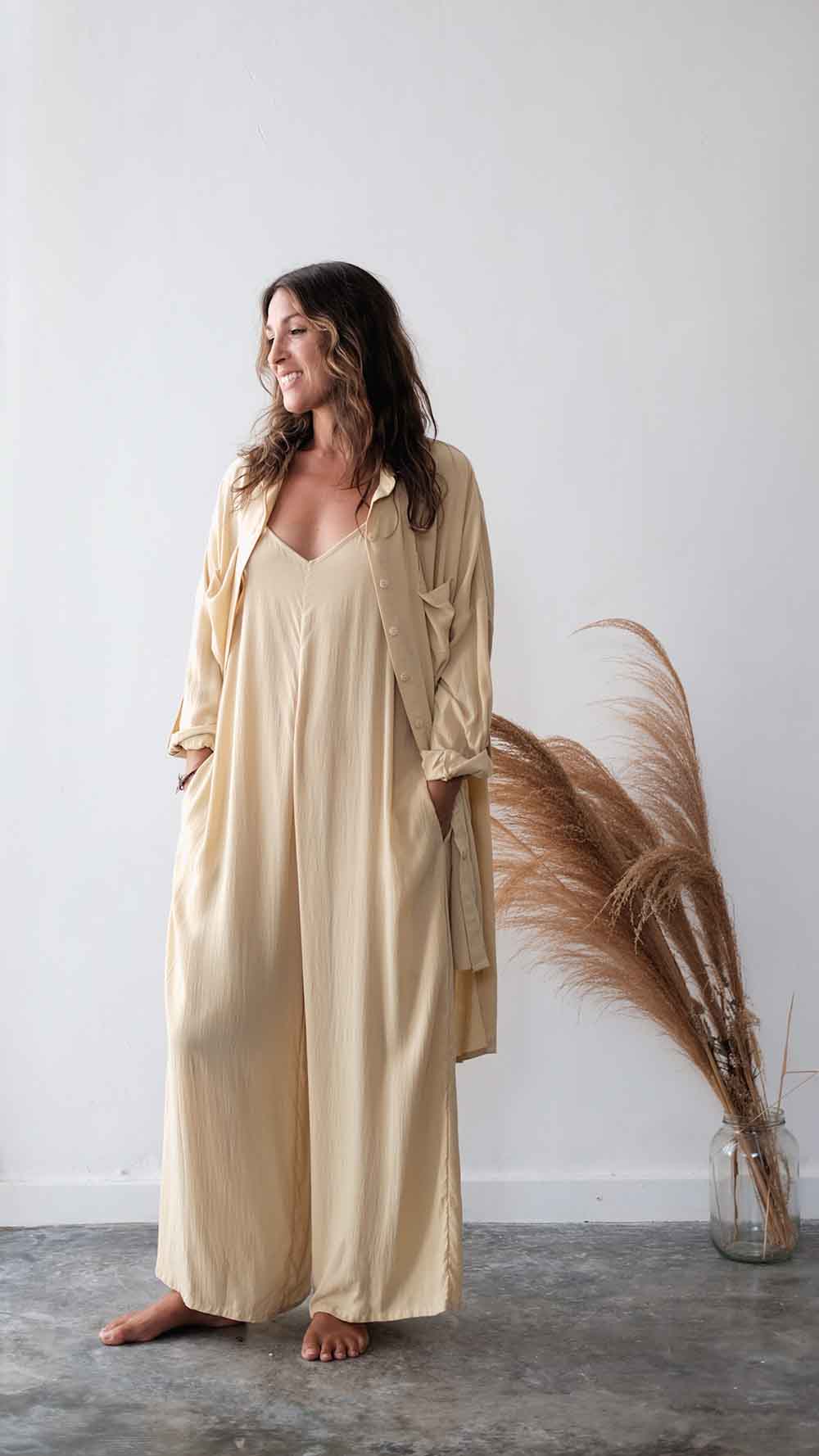 Woman wearing a beige outfit standing next to pampas grass against a white wall.