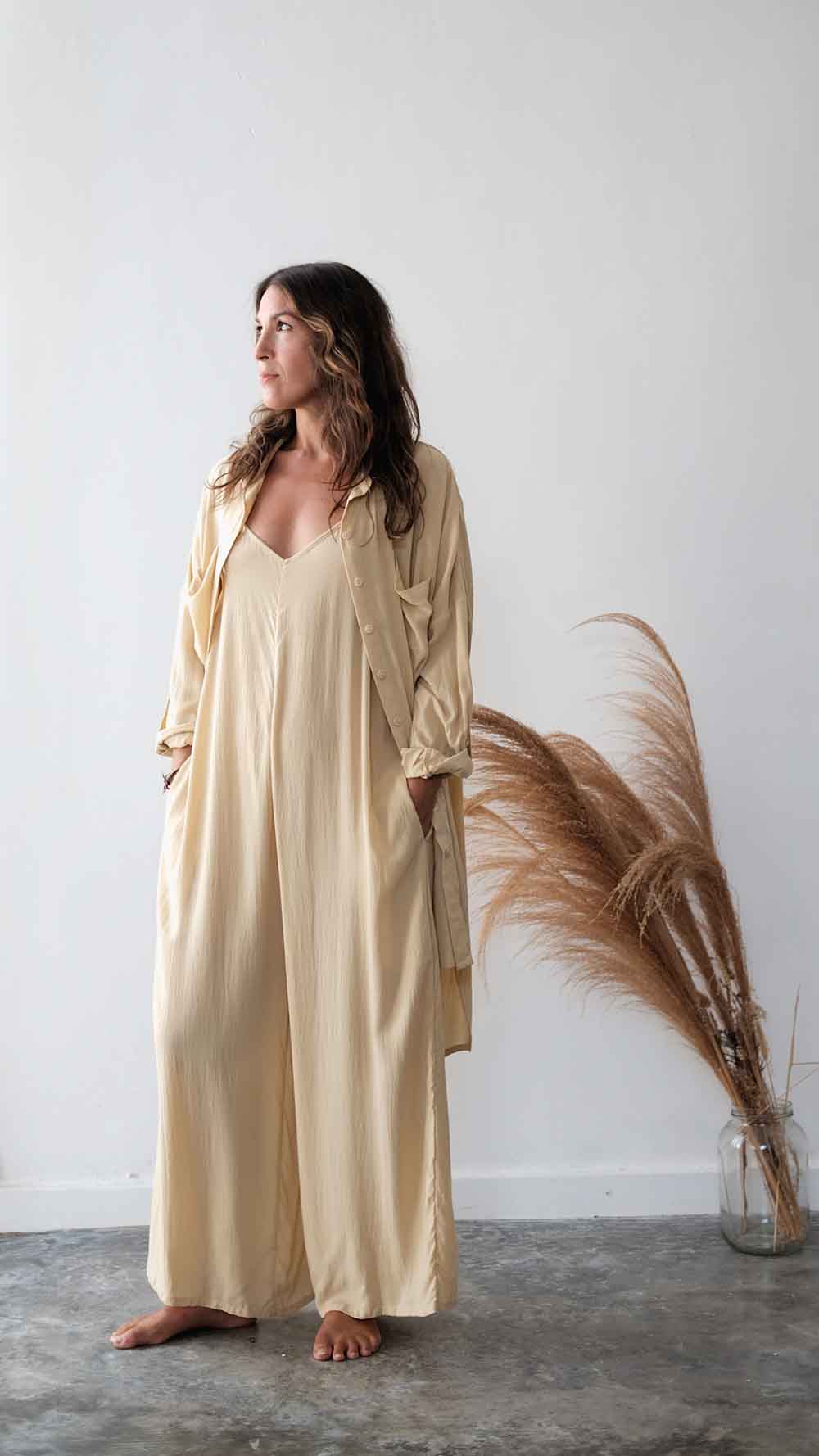 Woman wearing a beige long robe standing next to dried pampas grass against a white wall.