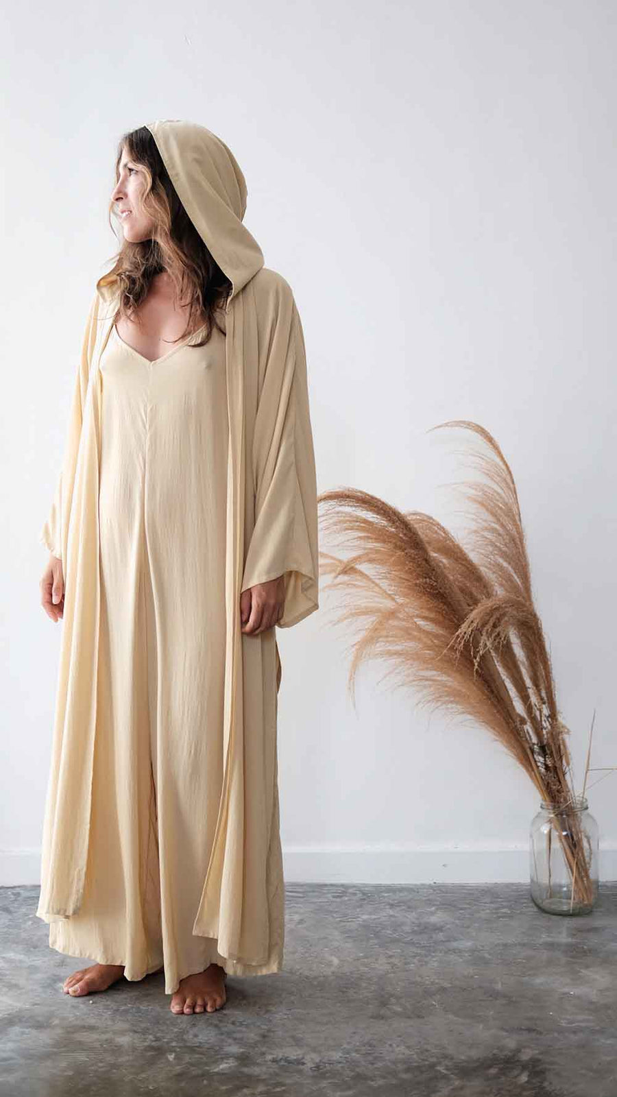 Woman wearing a long beige dress with a hood in a minimalistic setting.