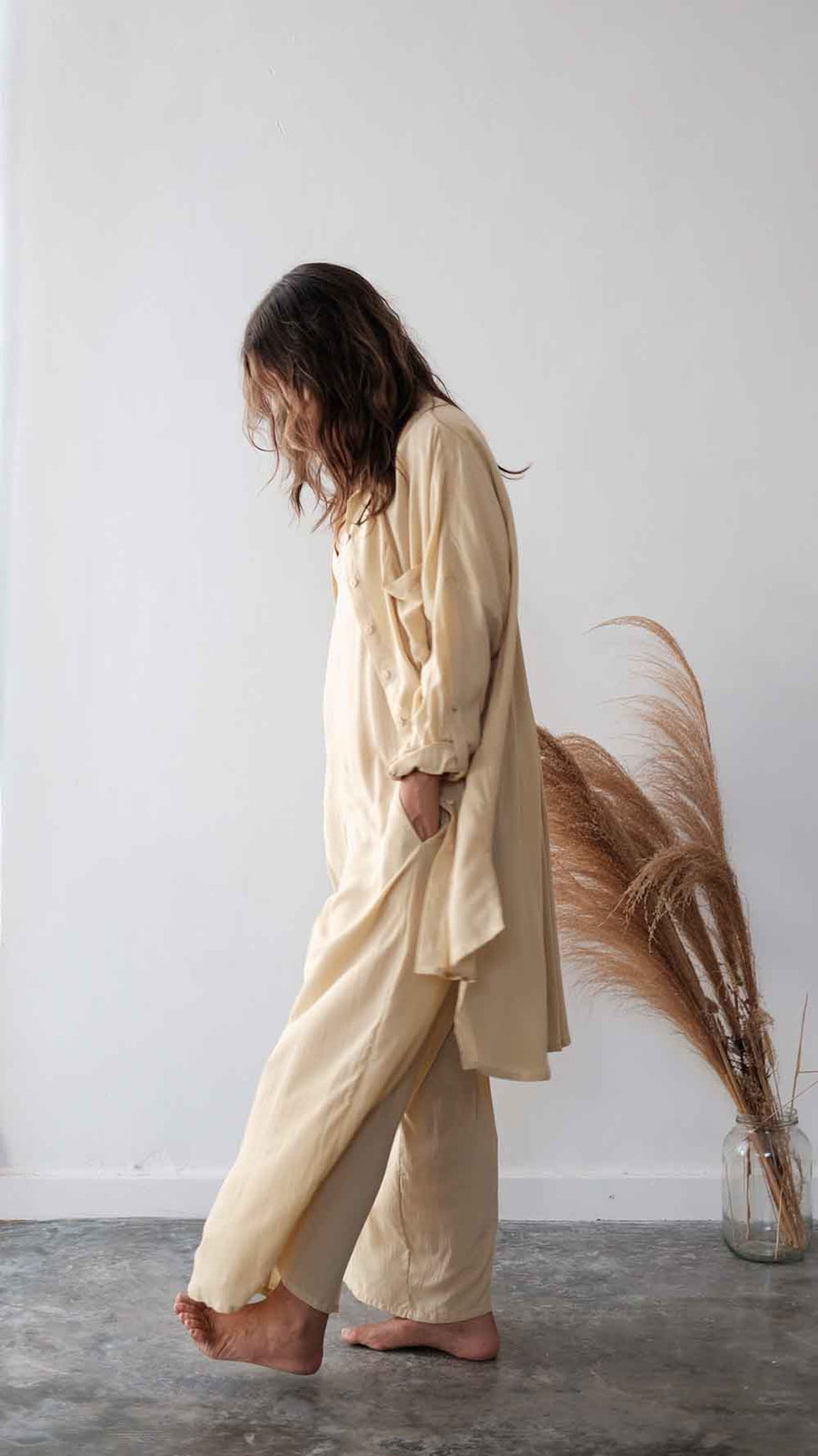 Person wearing a beige outfit standing in a minimalistic room with a vase of pampas grass.