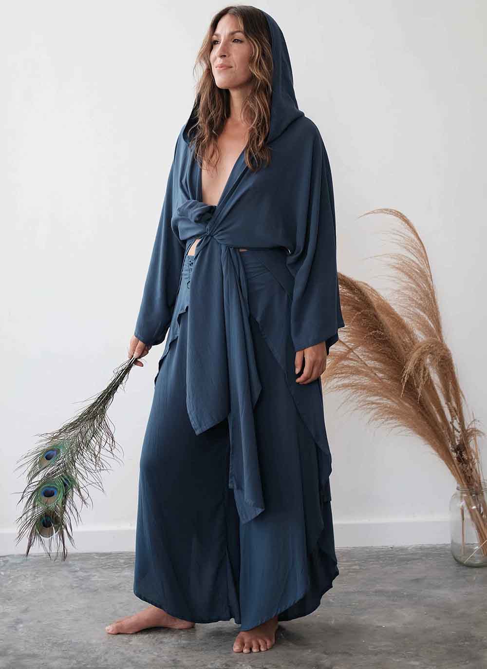Woman wearing a long, dark blue robe holding peacock feathers against a white wall.