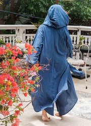 Person wearing a blue hooded garment standing outdoors with flowers and furniture in the background