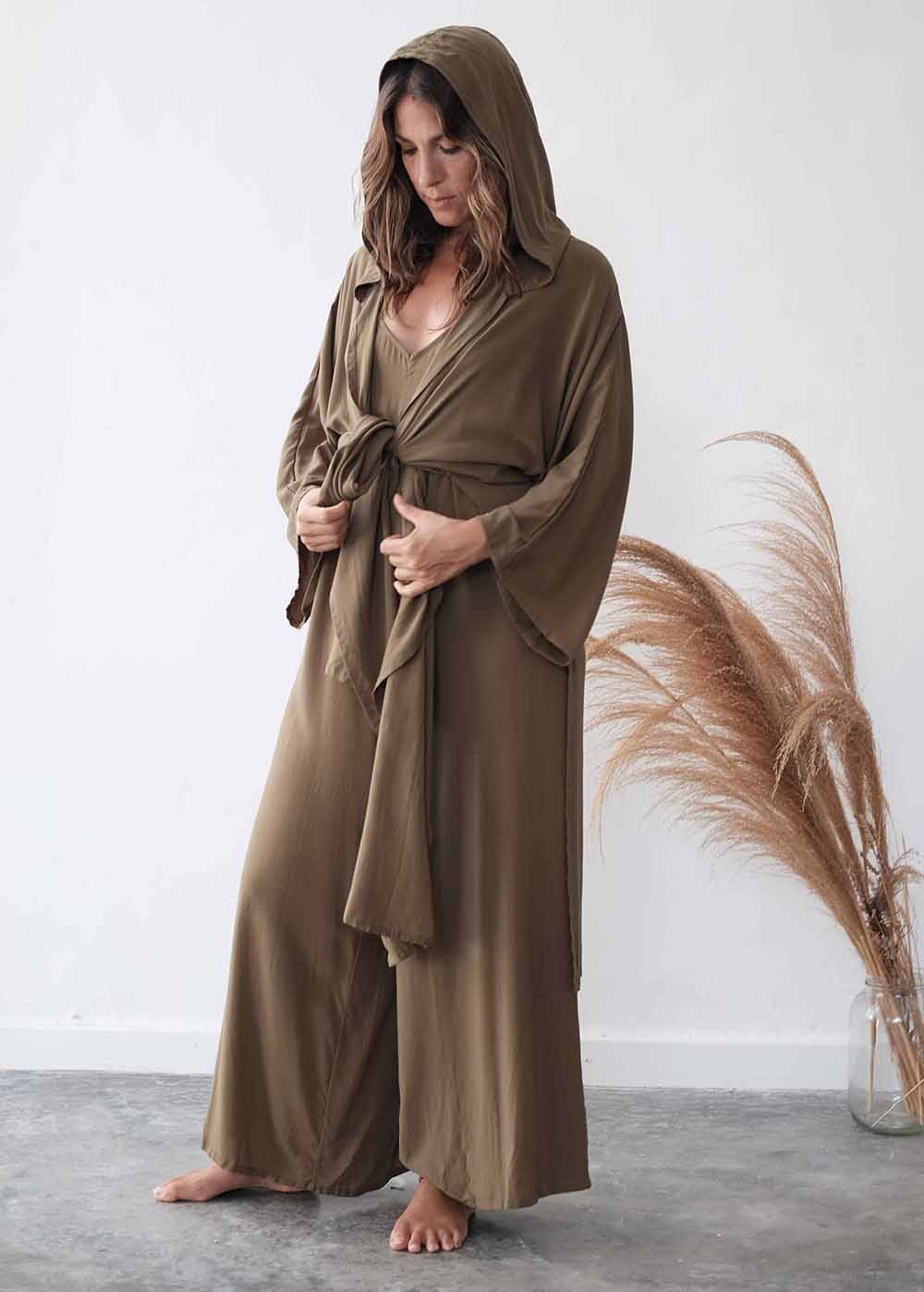 Woman wearing a long brown robe standing against a white wall with dried pampas grass.