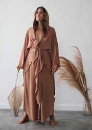 Woman in a long, light brown dress standing against a white wall with decorative elements.