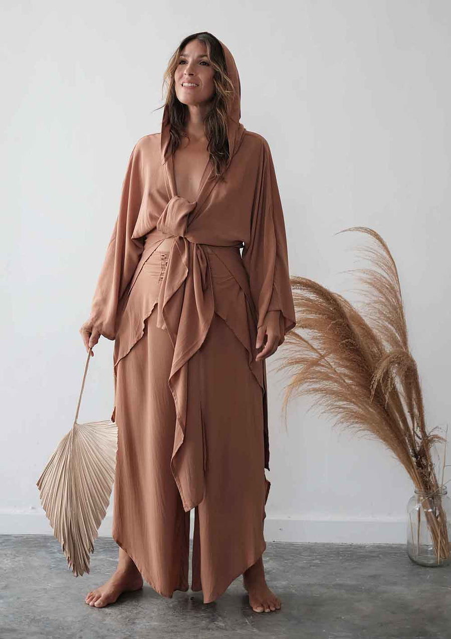Woman in a long, light brown dress standing against a white wall with decorative elements.