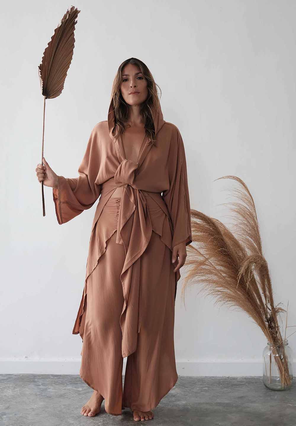 Woman in a long, light brown dress holding dried plants against a white wall.