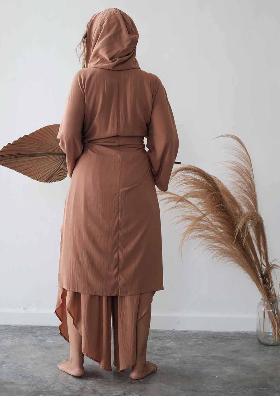 Person wearing a brown outfit with a hood, standing against a white wall with decorative elements.