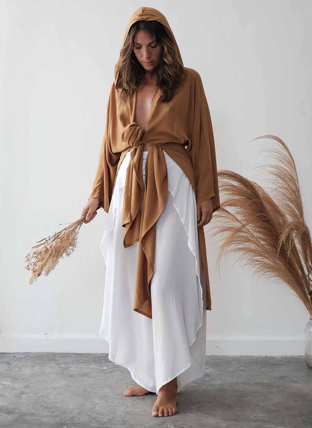 Woman wearing a brown top and white skirt holding dried pampas grass against a white wall.