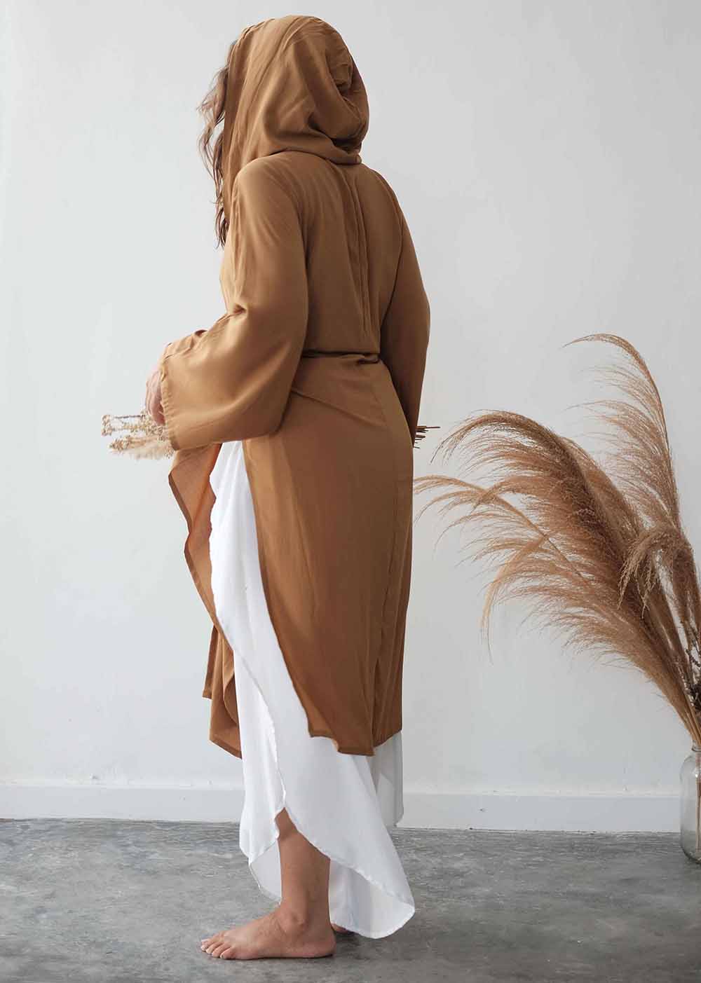 Person wearing a brown robe with a hood, standing against a white wall with decorative elements.