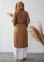 Person wearing a brown hijab and long brown coat with white pants, standing against a white wall with dried plants.