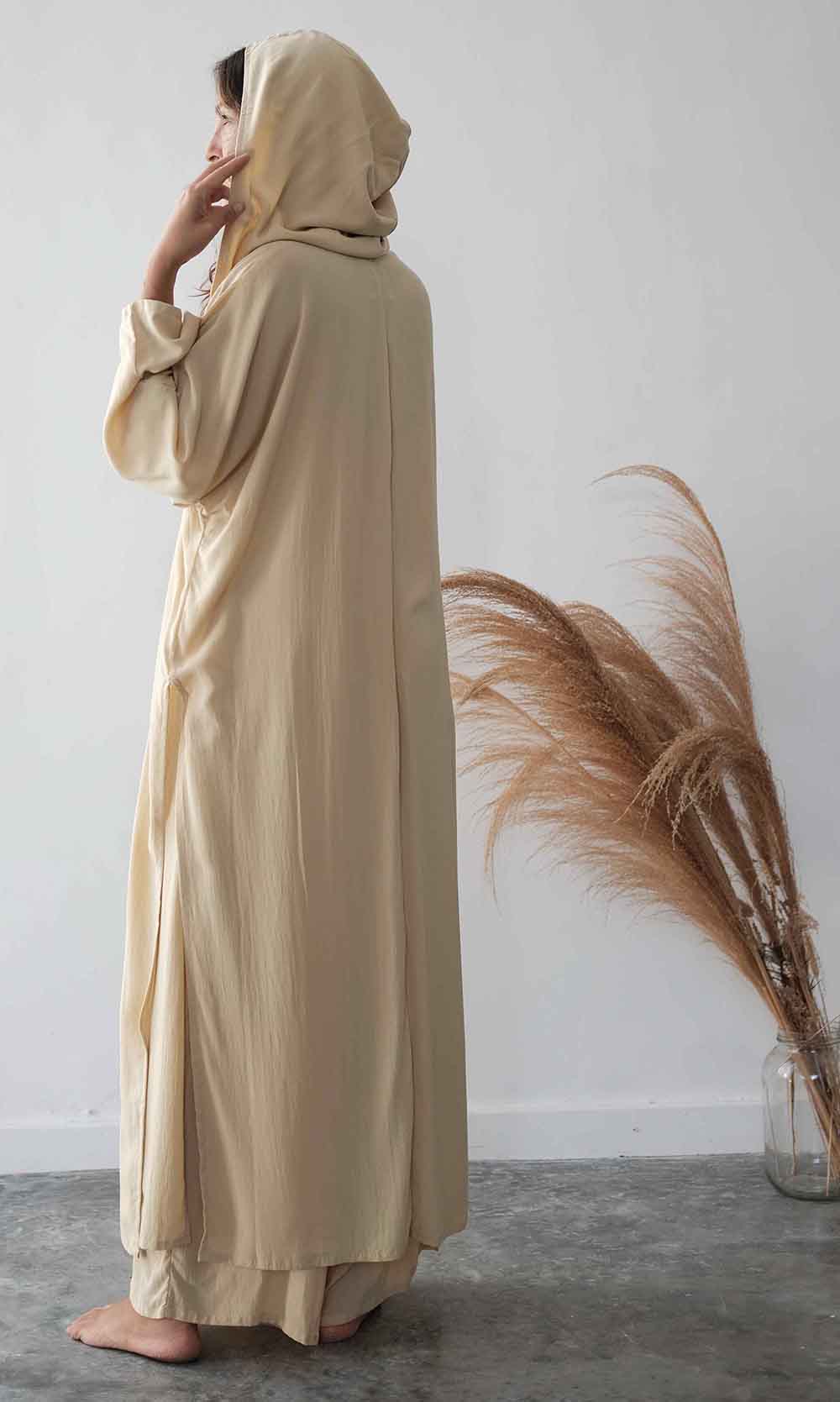 Woman wearing a beige abaya standing next to dried pampas grass against a white wall.