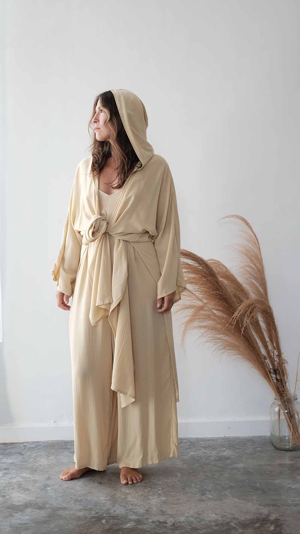 Woman wearing a beige hooded dress standing against a white wall with dried pampas grass.